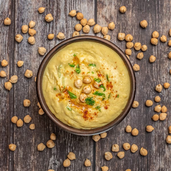 A bowl of hummus with chickpeas, paprika, and parsley, surrounded by scattered chickpeas on a wooden table.